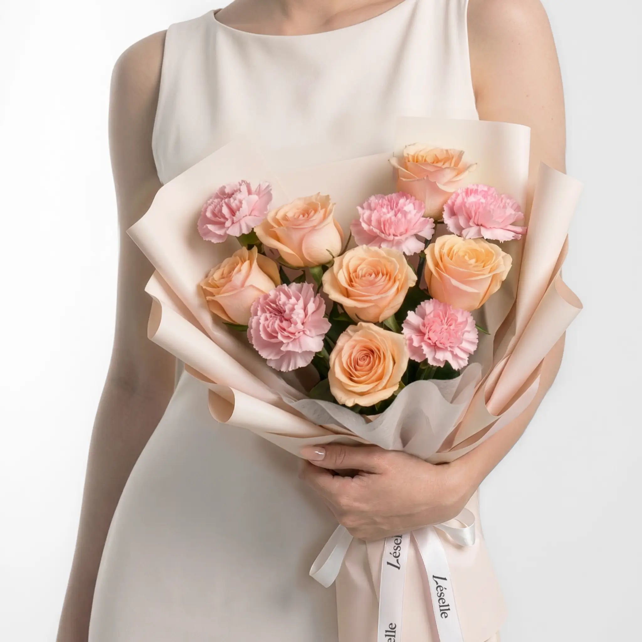Bouquet of champagne roses and blush carnations held by a person in a white dress on a light grey background